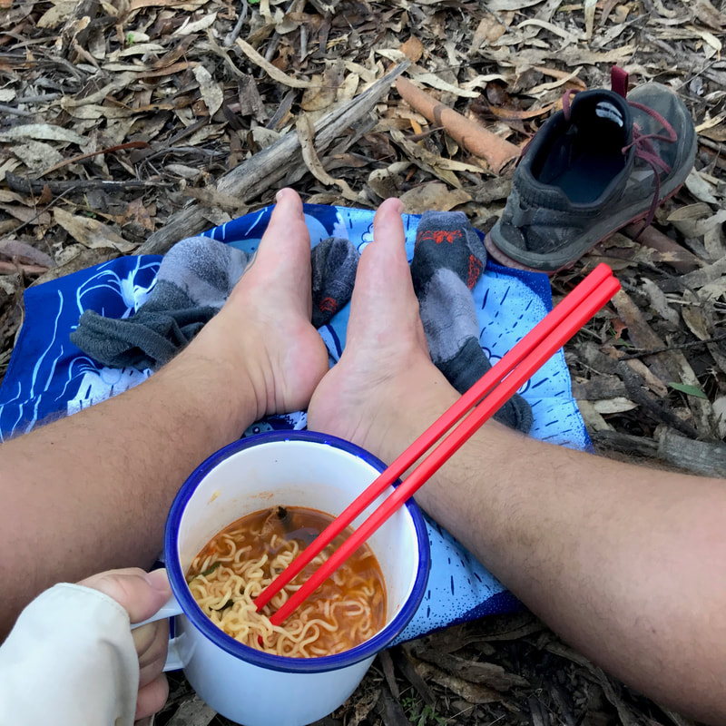 POV looking down at bare feet, a hand holding the cup of noodles, a blue bandana on the ground, shoes, socks and leaf litter.