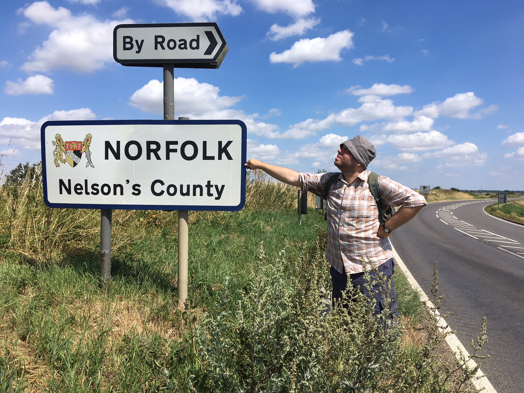 Person standing beside a sign saying NORFOLK Nelson's Country - and By Road