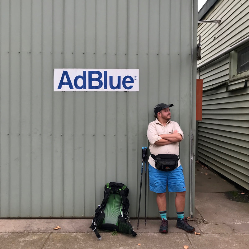 Person in front of a grey colorbond wall with a sign saying AdBlue on it. The person is in blue shorts, wears a large bum bag and has a back pack on the ground beside them.