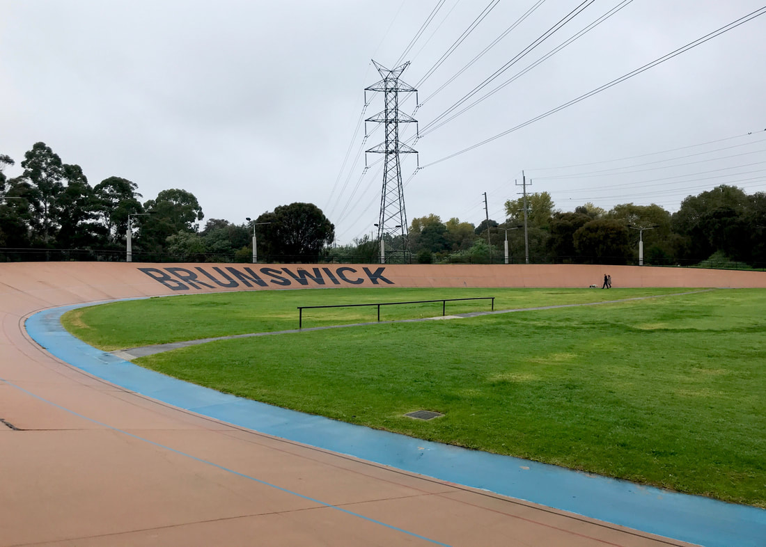 Outdoor velodrome with a terracotta coloured wall with BRUNSWICK in large black letters. The centre is green grass and a large pylon carries wires overhead.