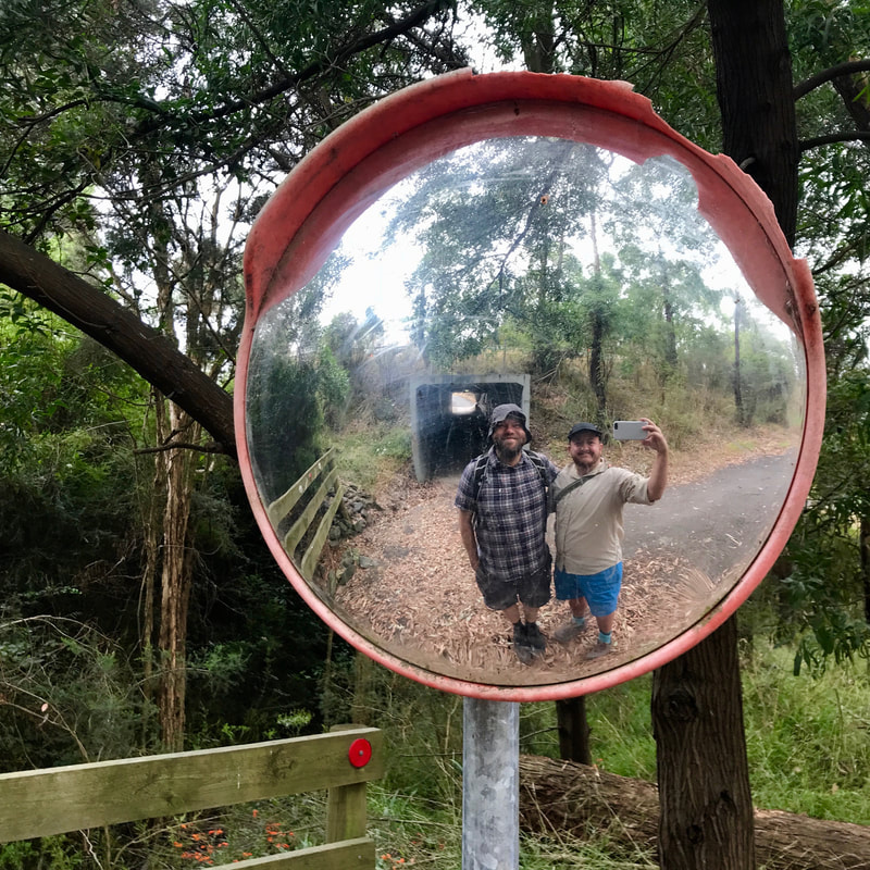 Two smiling people in a fisheye mirror with bush, trail and pedestrian tunnel behind