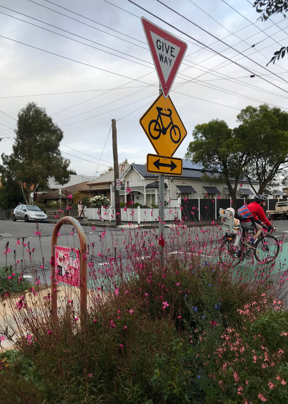 Pink flowers planted at a street corner below a bike and give way sign, in the background a person pedalling a bike with a young child on the back