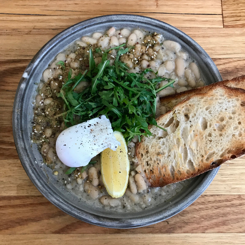 Grey bowl with pale brown beans topped with poached egg, rocket, toast and a slice of lemon