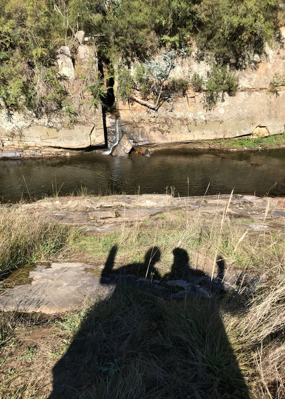 Shadow of two people waving, with the river behind. On the far side of the river is a narrow split in the rock face, from which a small trickle of water falls to the river