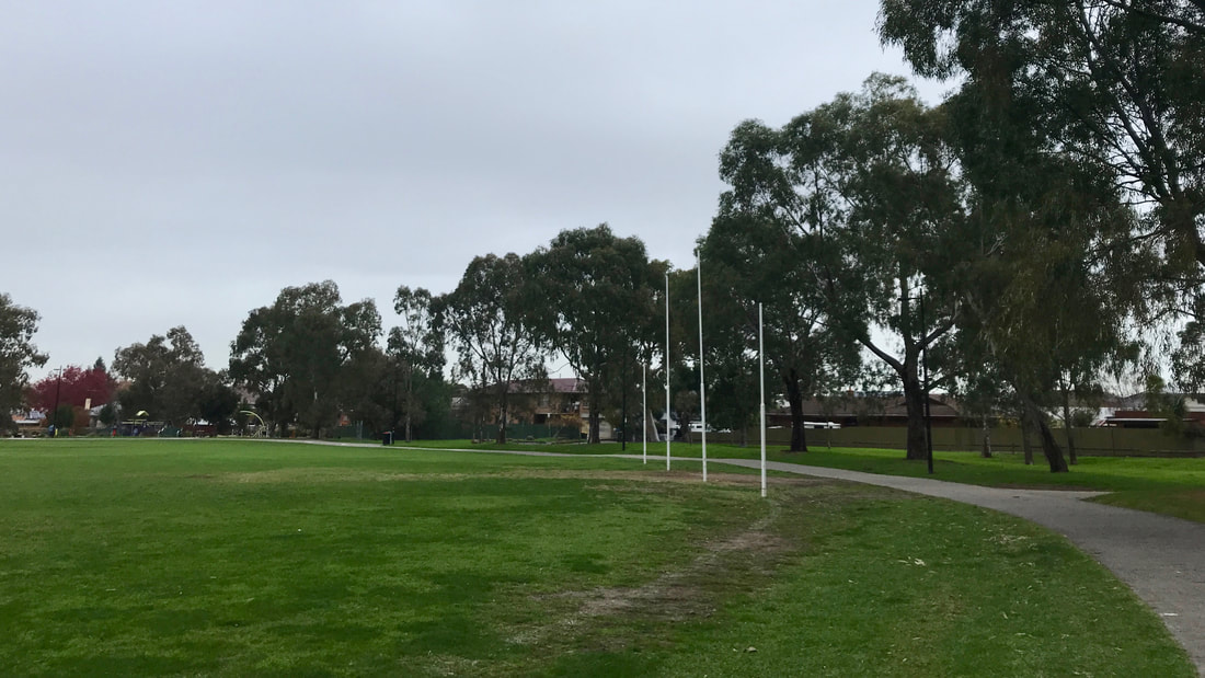 Concrete path and line of trees curving around a green oval with four white Australian Rules goal posts