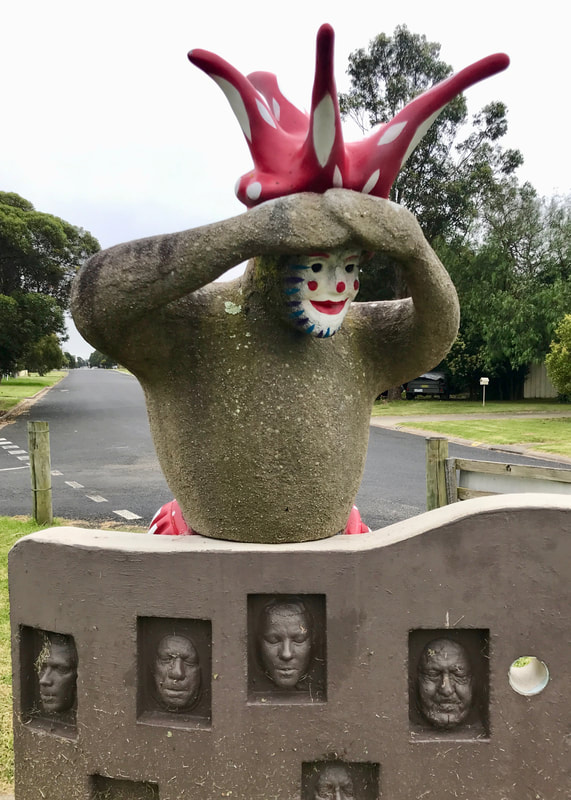 Grey concrete sculpture of a jester with a red and white painted hat, boldly painted face and the glimpse of red and white bloomers