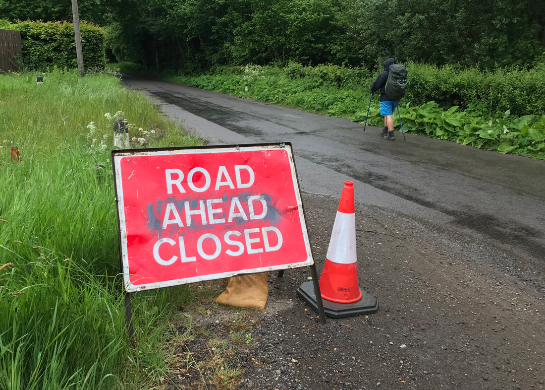 person walking down road with 'road ahead closed' sign