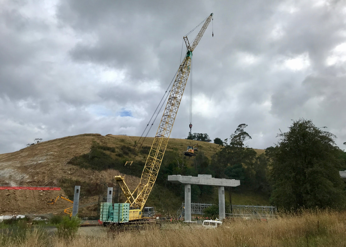 grassy hill under grey sky, a huge yellow crane lifts an excavator into a building site. the site contains the concrete piles of a future highway bridge.