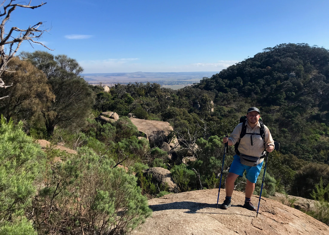 Person with blue shorts, bag and trekking poles standing on a large slab of granite that curves away to a view of hills and plains beyond