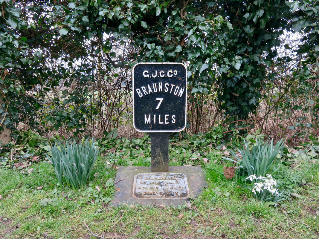 Painted steel sign saying BRAUNSTON 7 MILES surounded by flowers