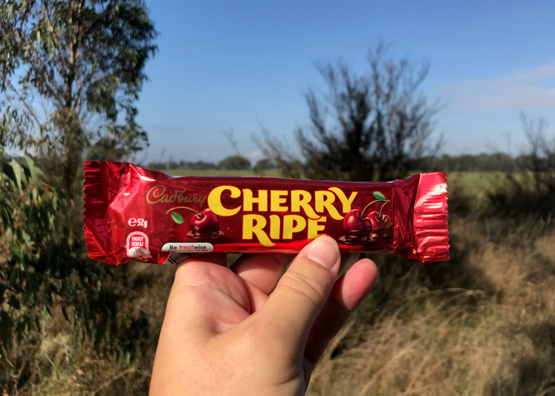 Close up of fingers holding a red wrapped Cherry Ripe chocolate bar, with trees and grasslands behind