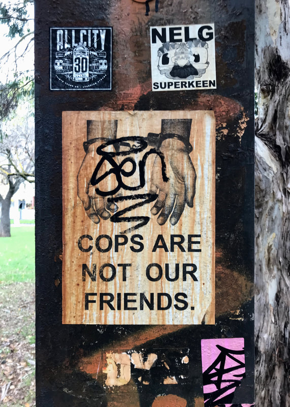 Steel pole with various stickers and signs, the main one showing hands in handcuffs and the text COPS ARE NOT OUR FRIENDS