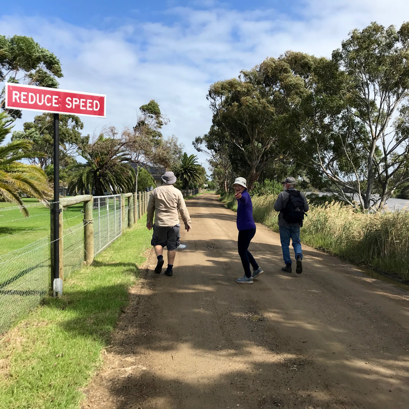 Three people walking away from the camera down a dirt road, the middle person turning back with a smile. They are beside a red sign saying REDUCE SPEED.