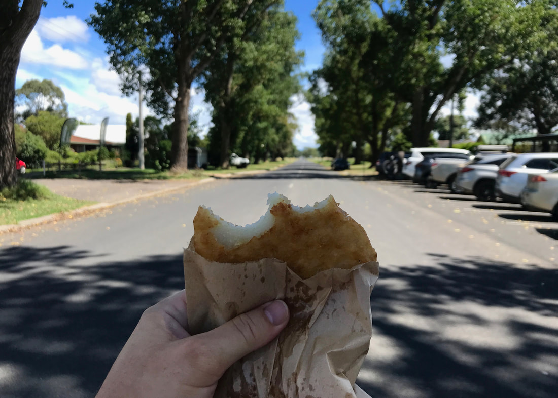 A hand holds a brown paper bag with a half-eaten potato cake, with an out of focus background of a long straght road between trees