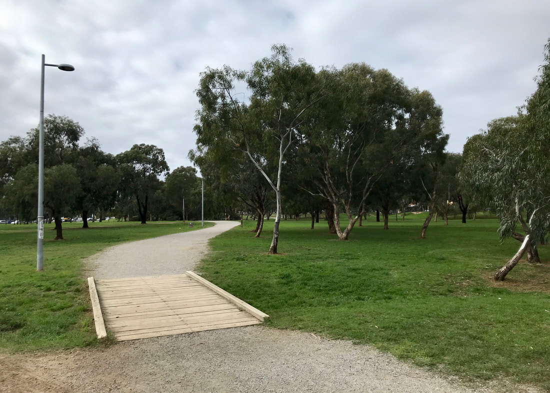 Gravel path and low wooden bridge through grassland dotted with eucalyptus trees
