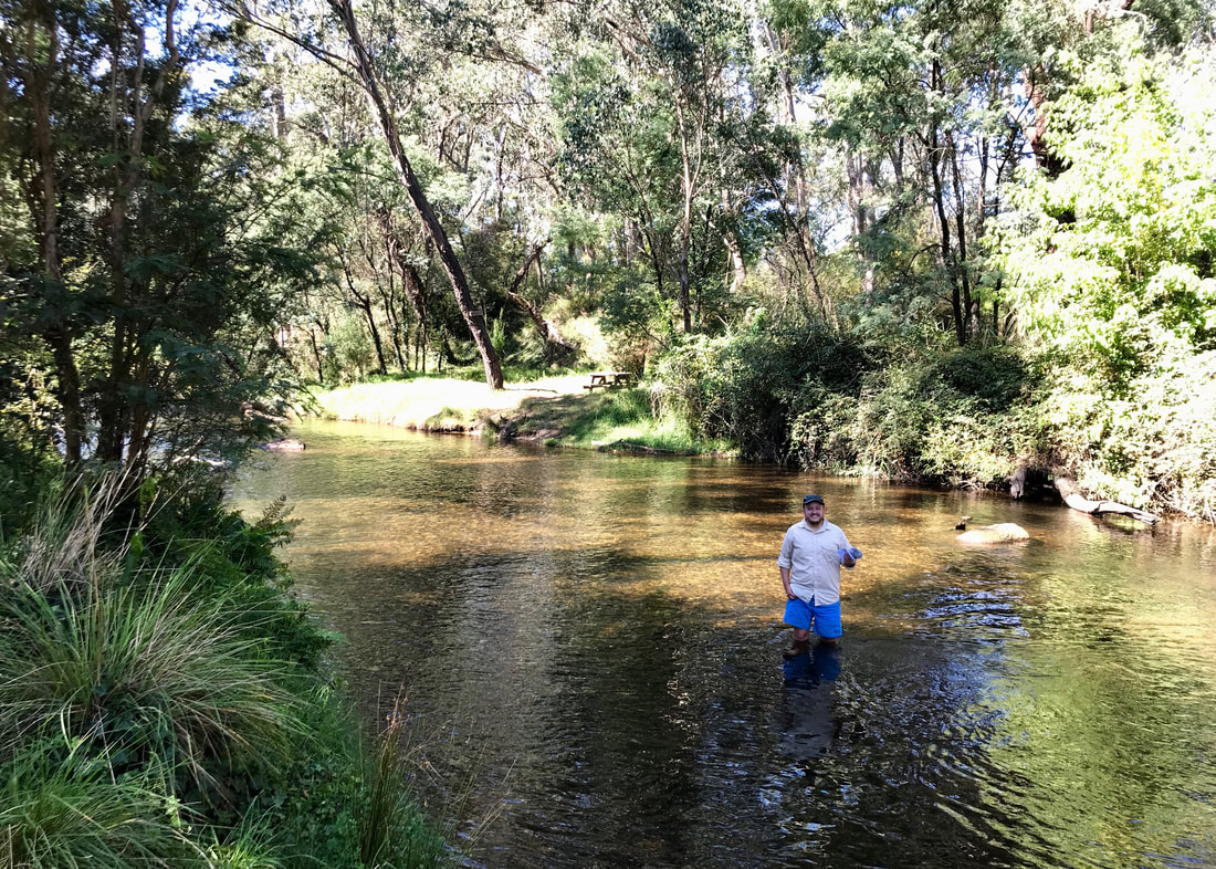 Smiling person in blue shorts standing knee-deep in a small river, with trees and grasses growing on each bank
