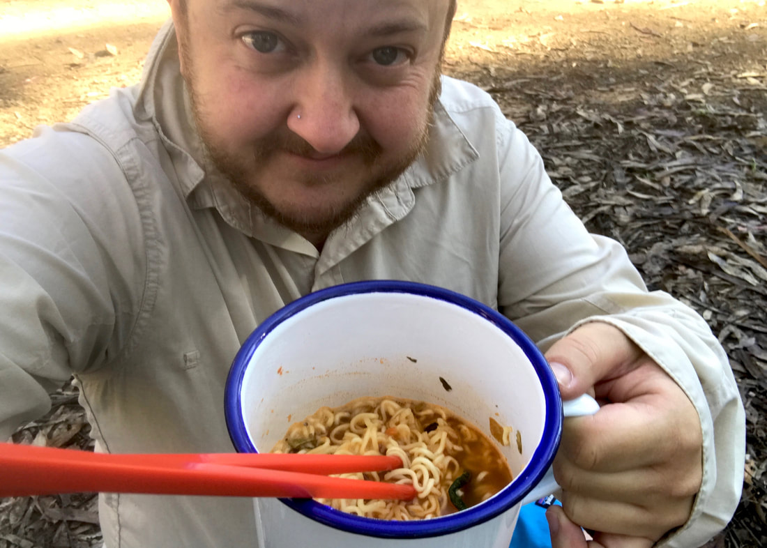 Selfie of person in beige shirt, holding a blue and white metal cup up to the camera. In the cup you can see noodles and a pair of red chopsticks.