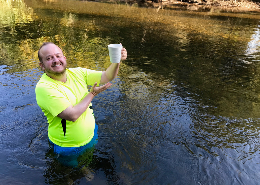 Person standing hip deep in a river, holding up a cup of tea and gesturing to it with a wide smile