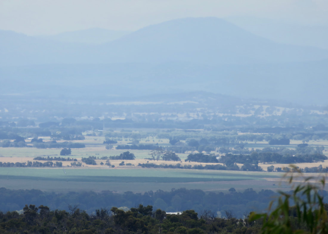 Hazy view over plains with paddocks and trees to blue hills beyond