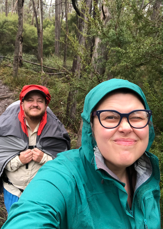 Selfie of two people in raincoats, walking on a bush path,smiling with squinting eyes