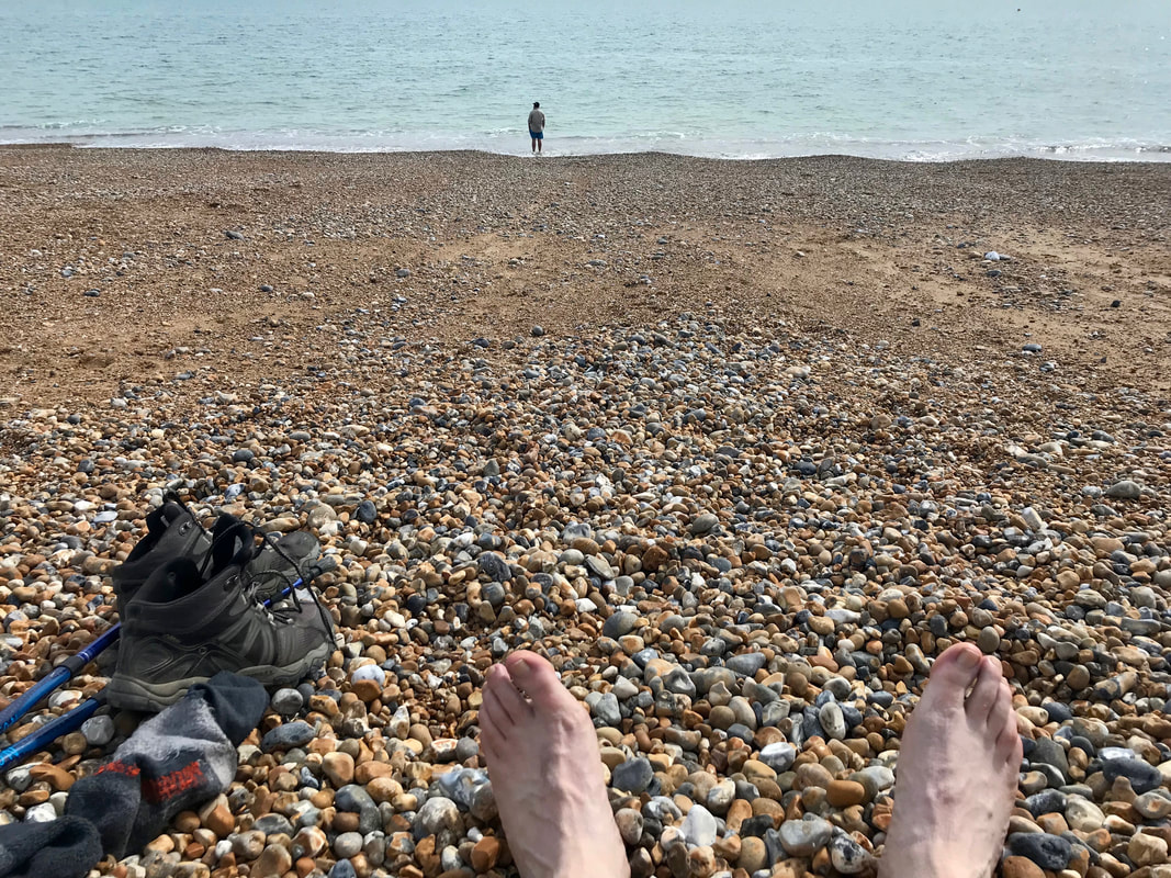 Feet, boots, socks and hiking poles and in the distance a person standing in the sea